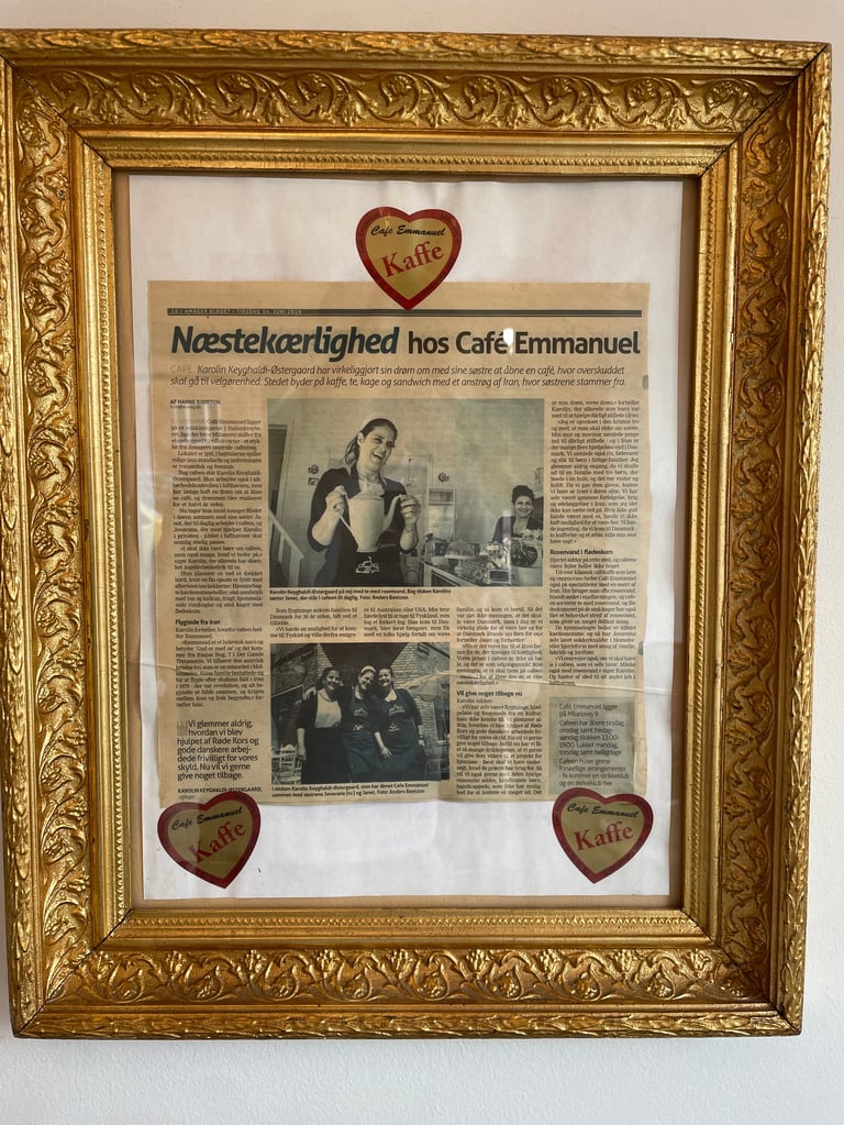 Framed newspaper article about Cafe Emmanuel in Danish, decorated with ornate gold frame and red heart-shaped coffee logos at corners